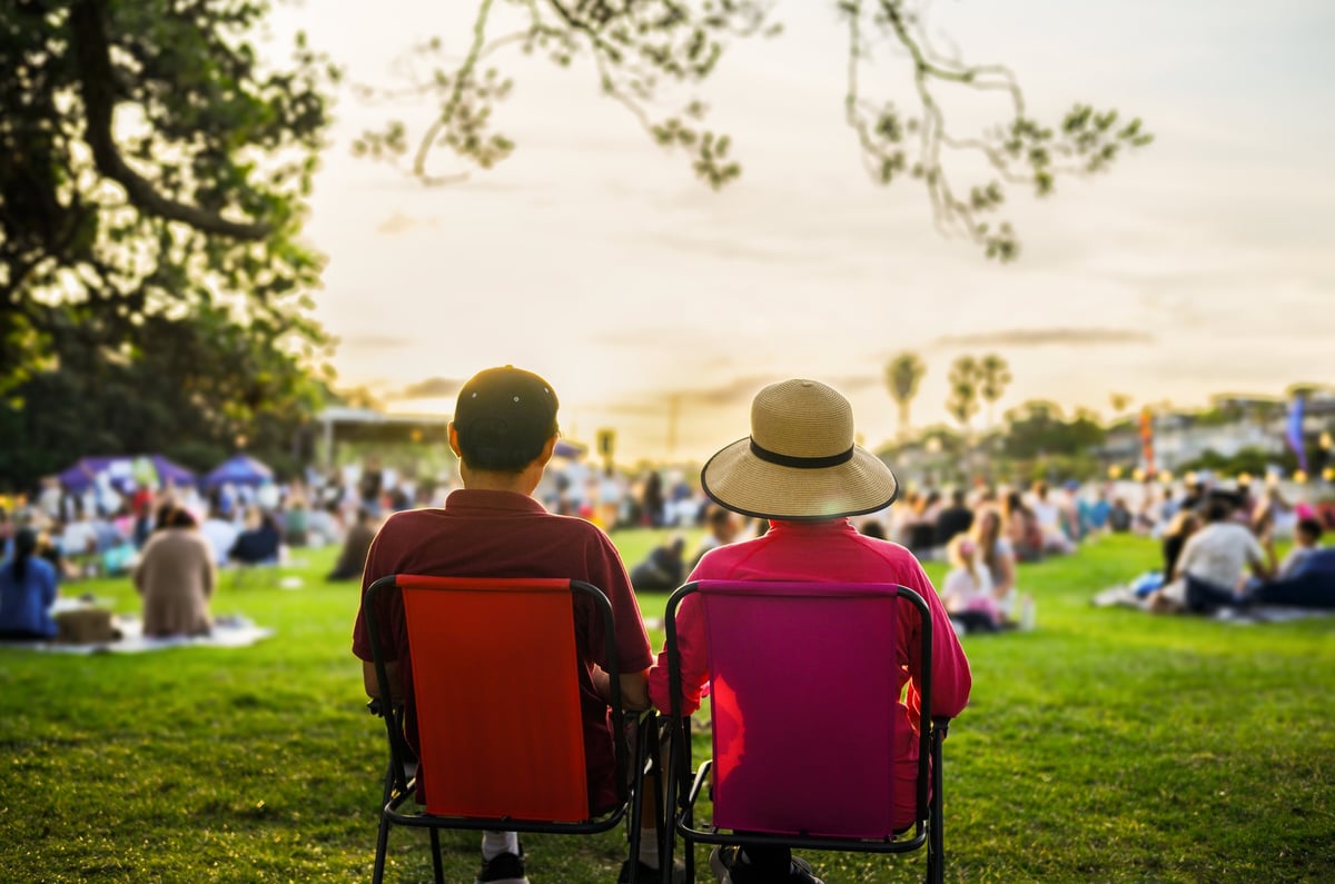 Couple sitting on the picnic chairs, listening to the music concert. Unrecognizable crowd of people in the park.